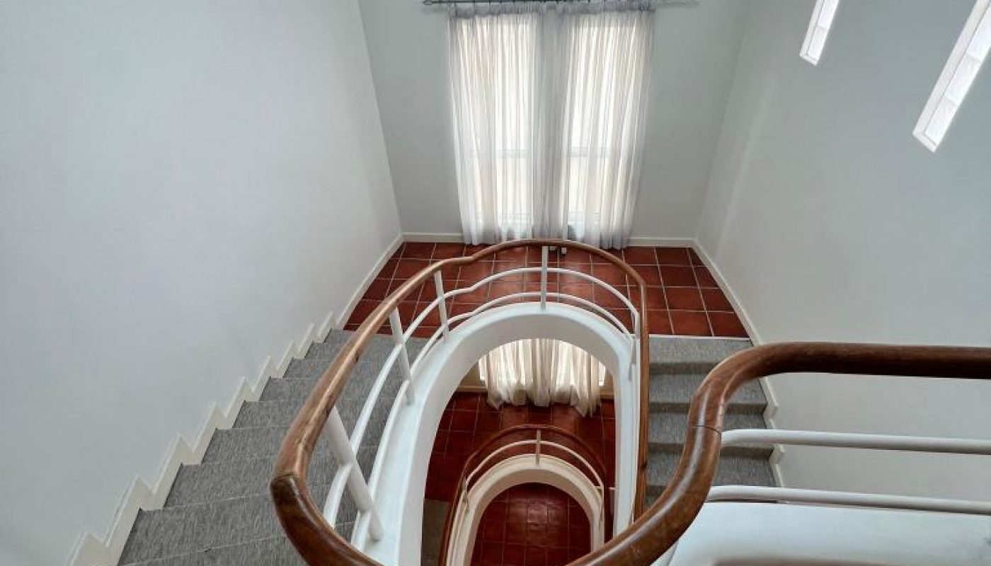 A spiral staircase with white railings and wooden handrail leads to a tiled floor, next to tall windows with sheer curtains letting in natural light.