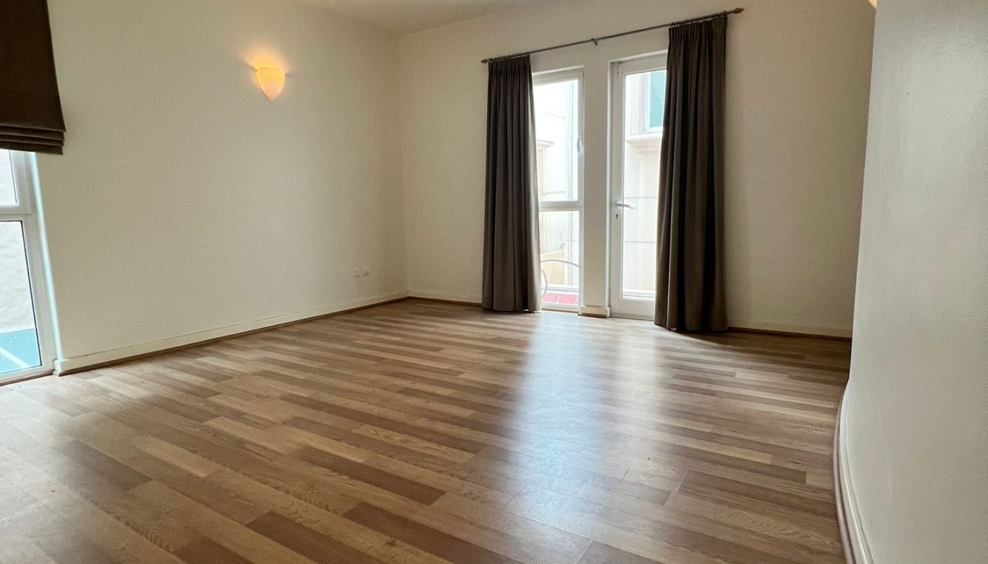 Empty room with wood flooring, white walls, a ceiling fan, two wall lights, and large windows with dark curtains letting in natural light.