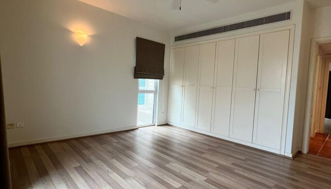 Empty room with light wood flooring, built-in white wardrobes, a ceiling fan, a small window with a dark shade, and a wall light.