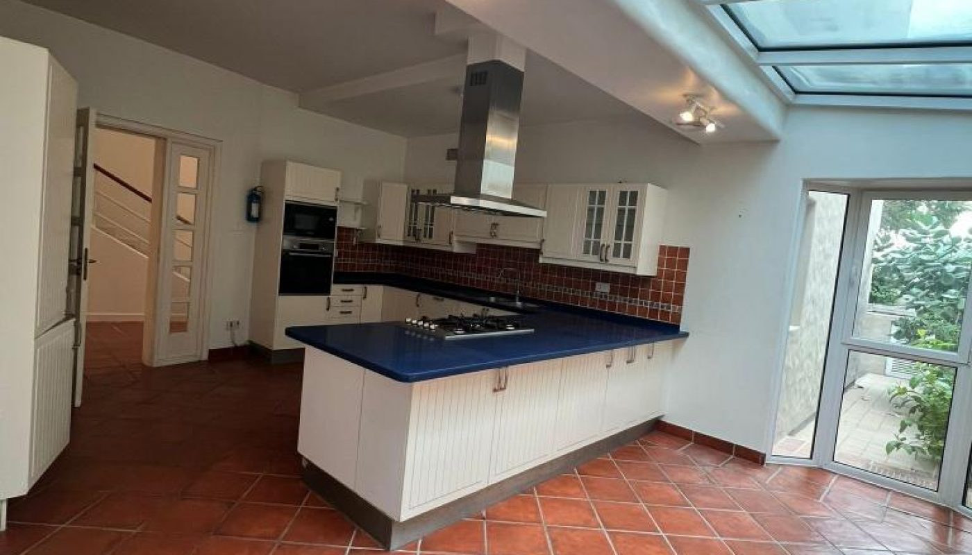 Spacious kitchen with terracotta tile flooring, central island with stovetop, white cabinets, skylights, and a glass door leading outside.