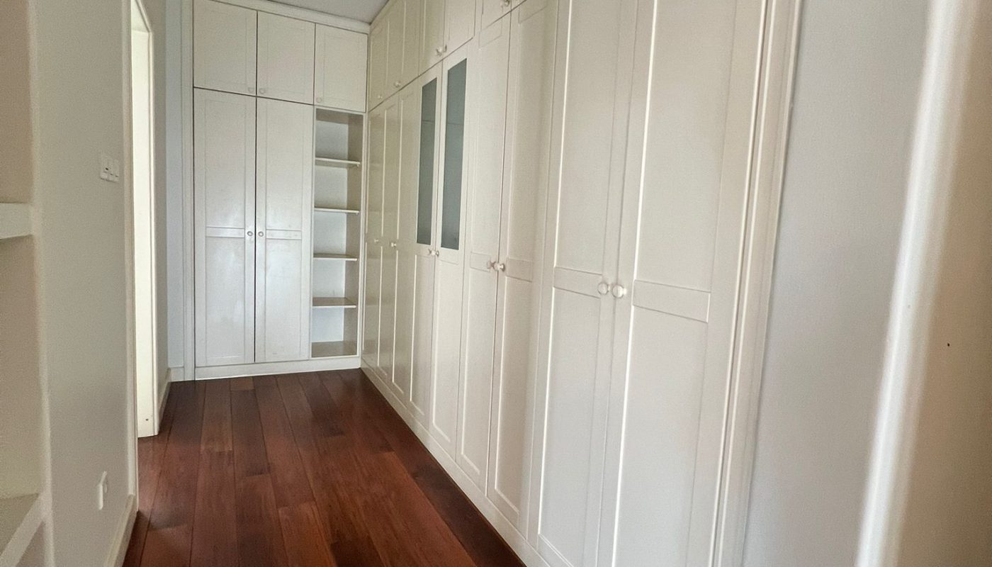 A hallway with wooden flooring, built-in white cabinets along the right wall, and shelves at the far end near a doorway.