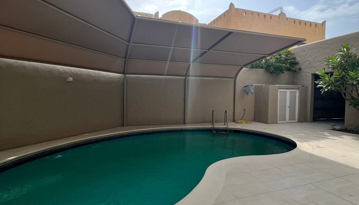 Oval-shaped swimming pool with clear water, surrounded by a tiled deck and shaded by a large canopy in a walled outdoor area under a partly cloudy sky.