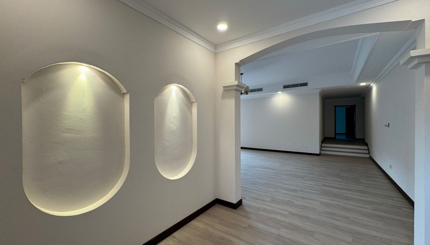 Spacious empty room with light wood flooring, two recessed wall niches with spotlights, white walls, and an arched entryway leading to another area.