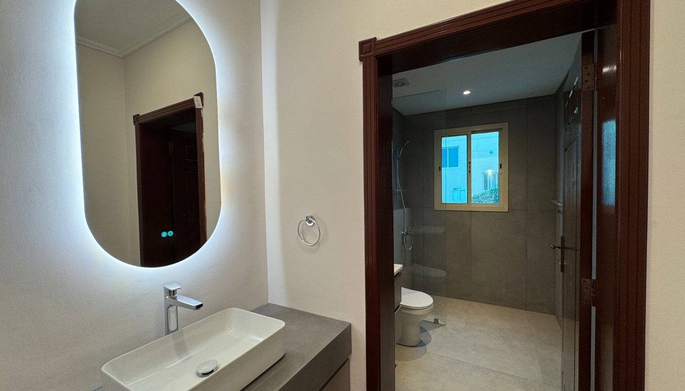 Modern bathroom with a rectangular sink, illuminated oval mirror, and a view into a shower and toilet area through a wooden-framed doorway.