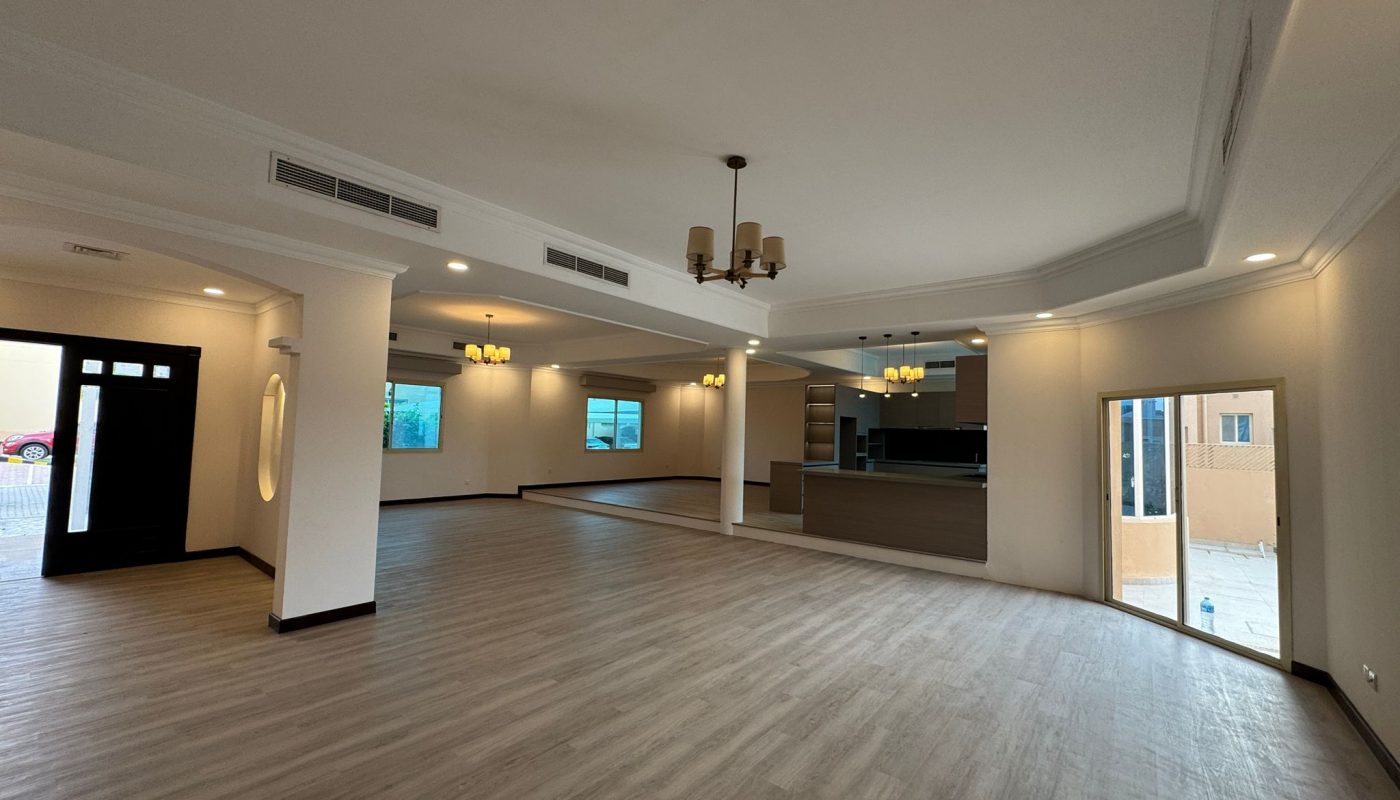 Spacious empty room with light wood flooring, white walls, ceiling lights, large windows, and an open doorway leading outside.