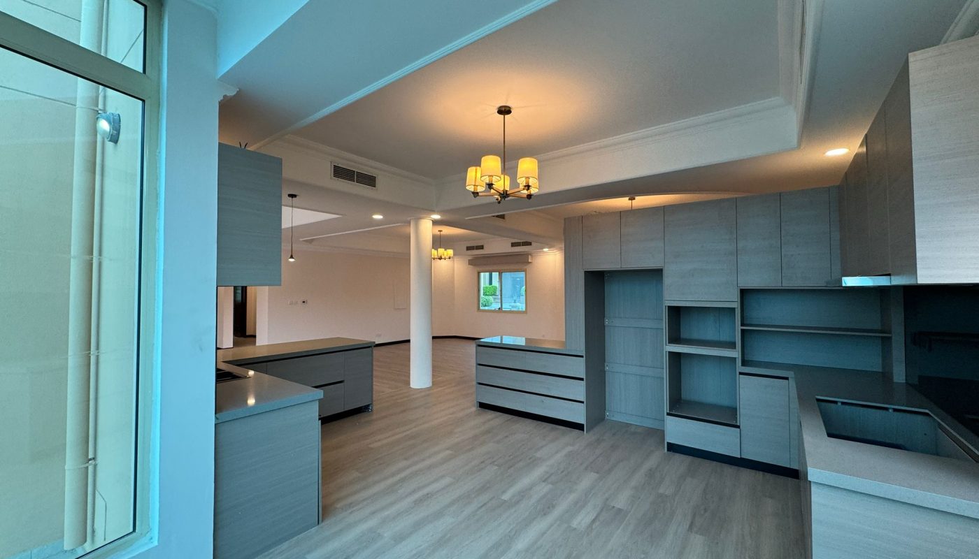 Spacious modern kitchen with light wood cabinets, large windows, and open layout connected to a living area. Ceiling lights and natural light brighten the space.