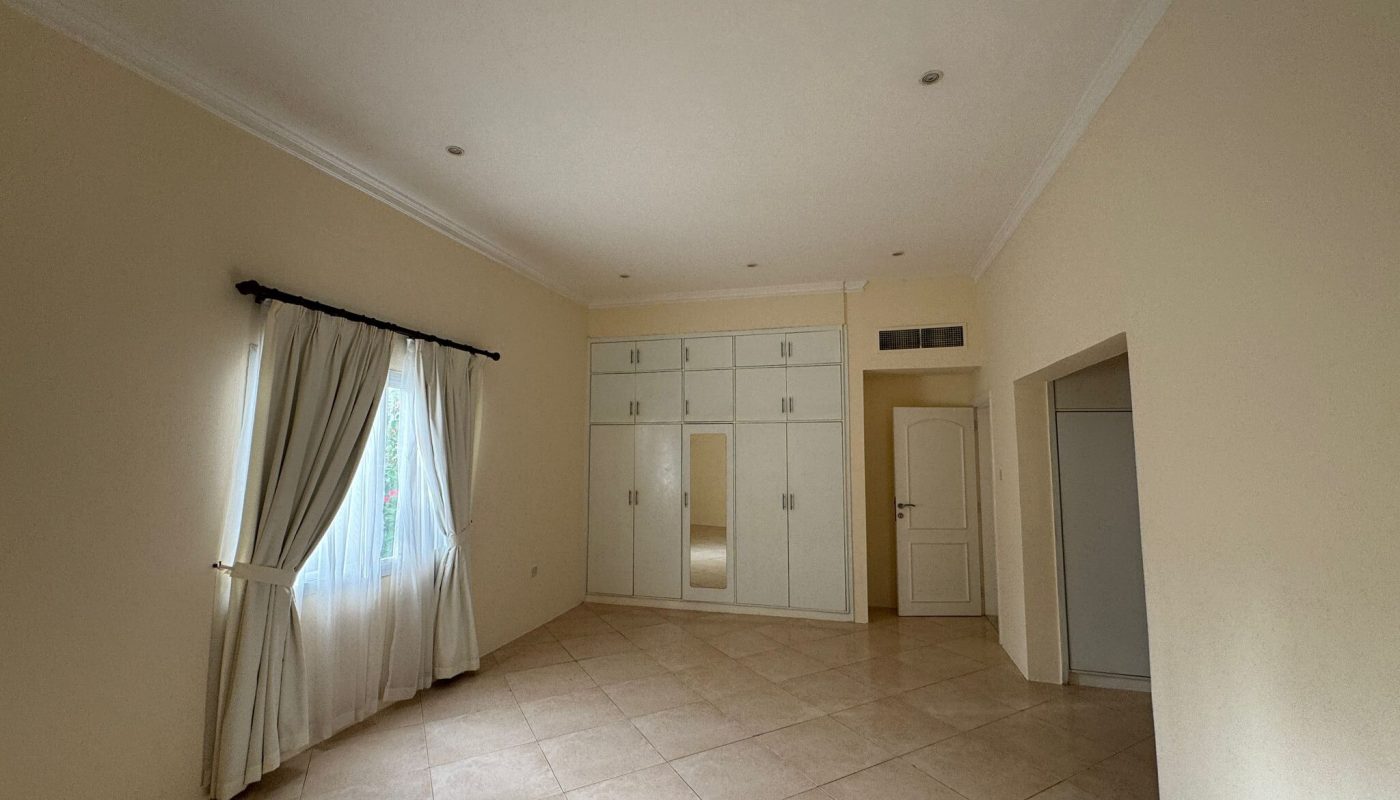 Spacious empty room with tiled floor, cream walls, a window with white curtains, built-in white wardrobes, and a closed door.