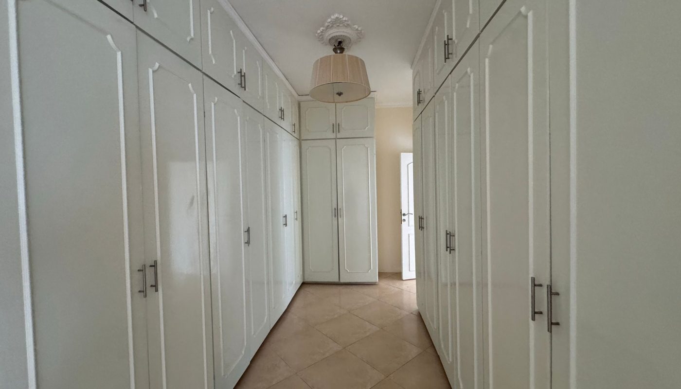 A narrow hallway with cream-colored cabinets on both sides, tiled floor, a ceiling light fixture, and a door at the end.