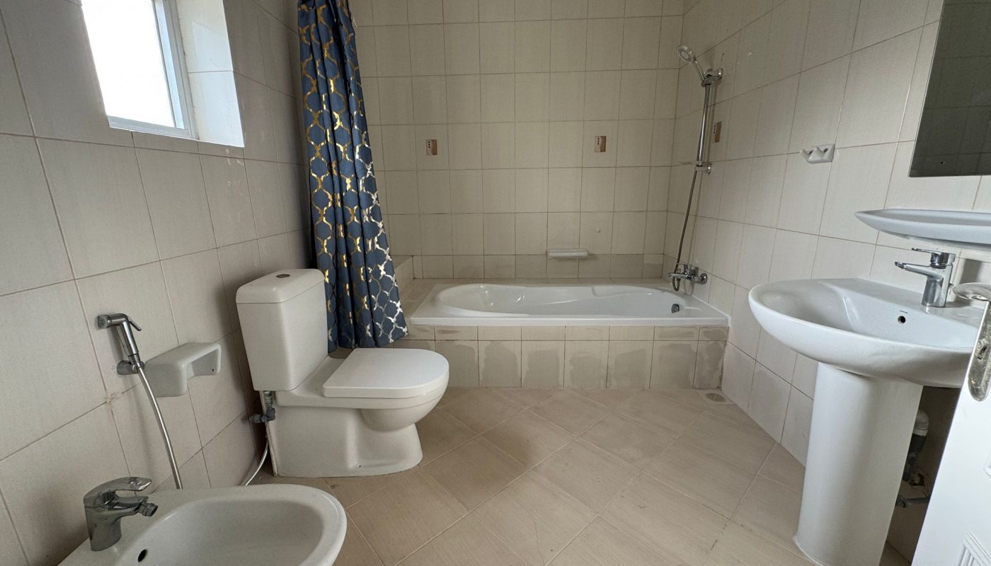 A bathroom with a bathtub, shower, toilet, bidet, sink, and a blue patterned shower curtain, all set against beige tiled walls and floor.