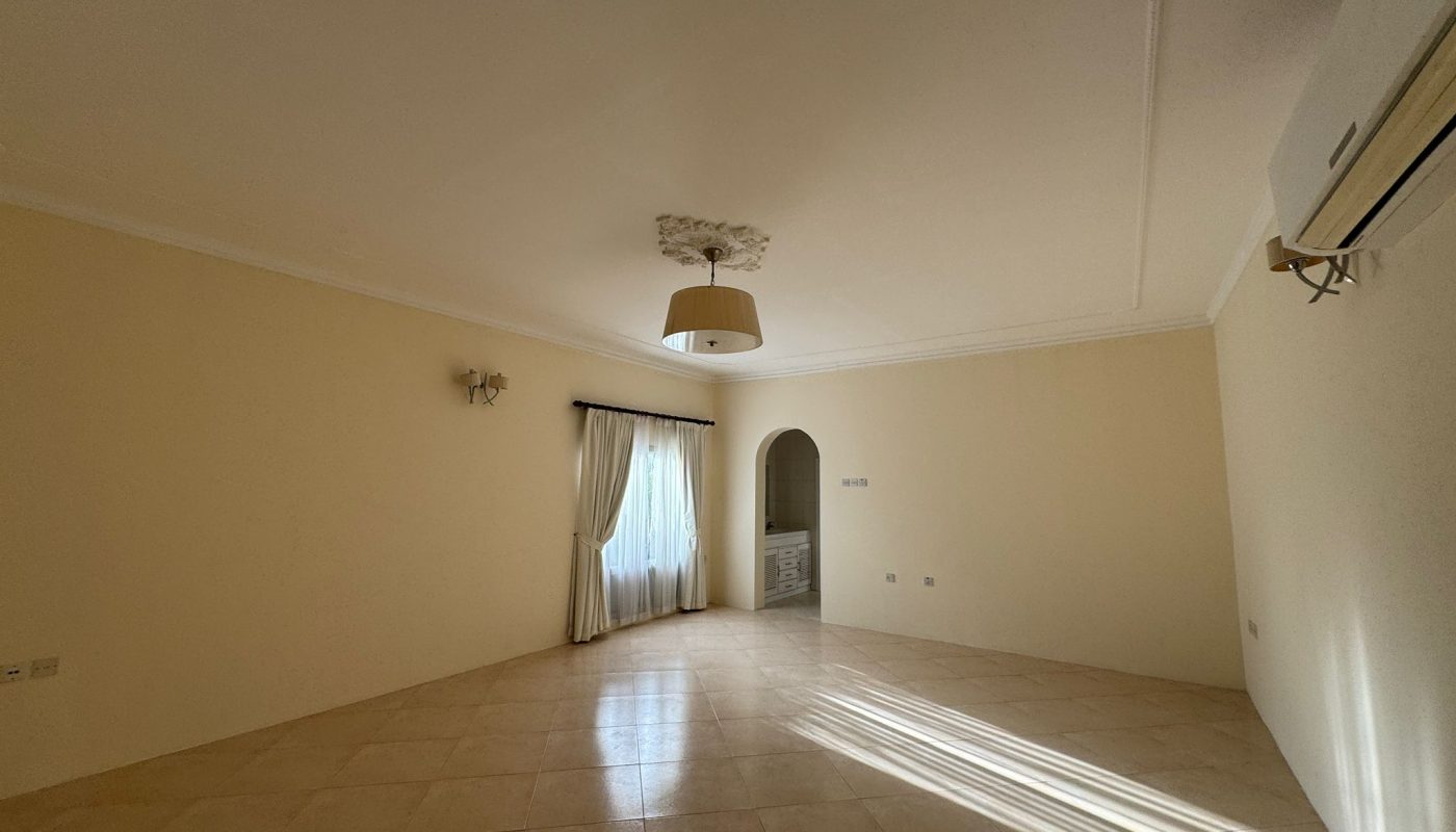 Spacious empty room with beige tiled floor, cream walls, a window with curtains, an air conditioner, and a ceiling light fixture. Sunlight streams in through the window.