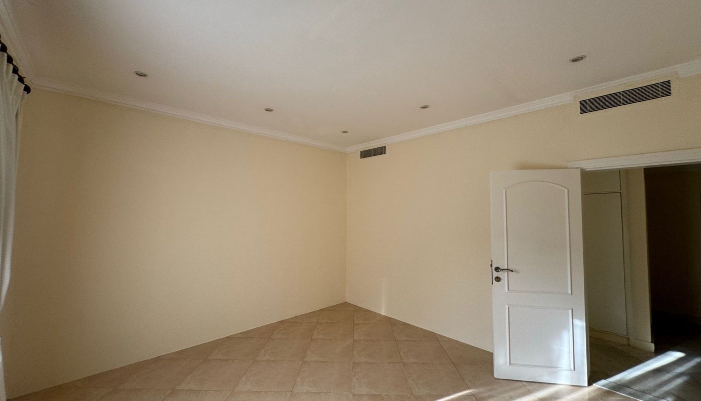 Empty room with cream-colored walls, tiled floor, white ceiling with recessed lights, an open white door, and sunlight streaming through a window.