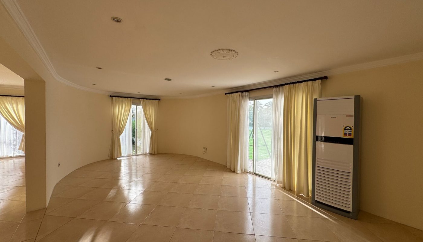 Spacious, empty living room with tiled floor, cream-colored walls, large windows with light curtains, and a standing air conditioner.