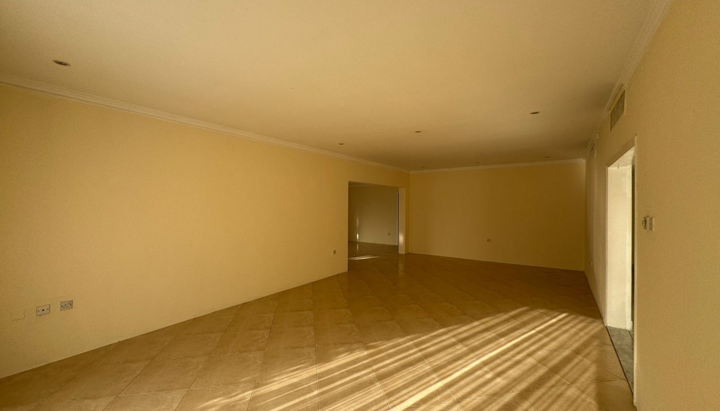 Empty room with beige tiled floor, cream-colored walls, and sunlight casting shadows through a window on the right. There are recessed ceiling lights and an open doorway at the far end.