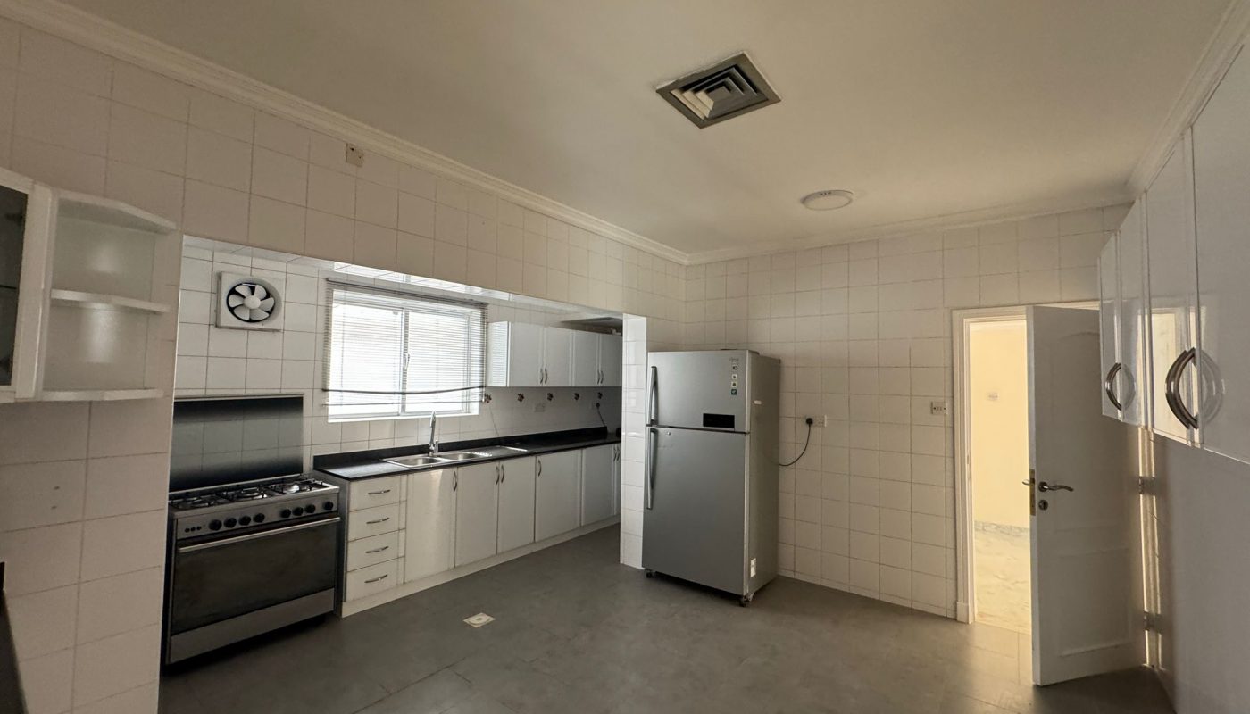 Spacious kitchen with white cabinets, stainless steel stove, refrigerator, wall clock, and tiled walls and floor. Door is open letting in natural light.