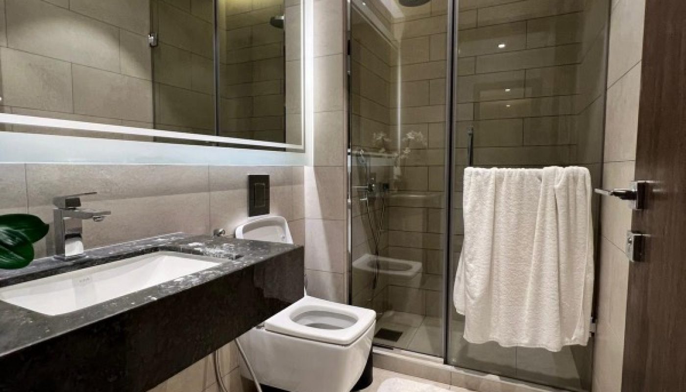 Modern bathroom with a sink, wall-mounted toilet, glass shower enclosure, white towel on the door, and beige tile walls and floor.