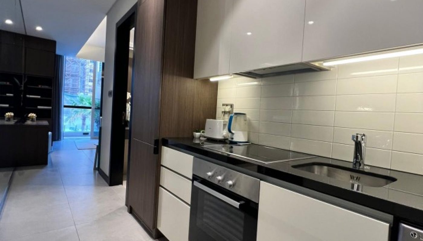 Modern kitchen with white cabinets, black countertop, built-in oven, sink, and backsplash tiles. Large window and dining area visible in the background.