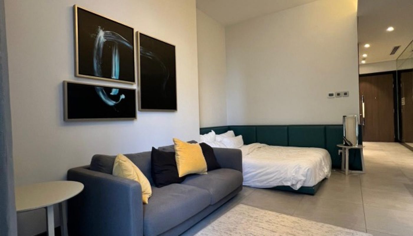 Modern studio apartment with a gray sofa, white and yellow pillows, bed with green headboard, abstract wall art, side table, rug, and pendant light.