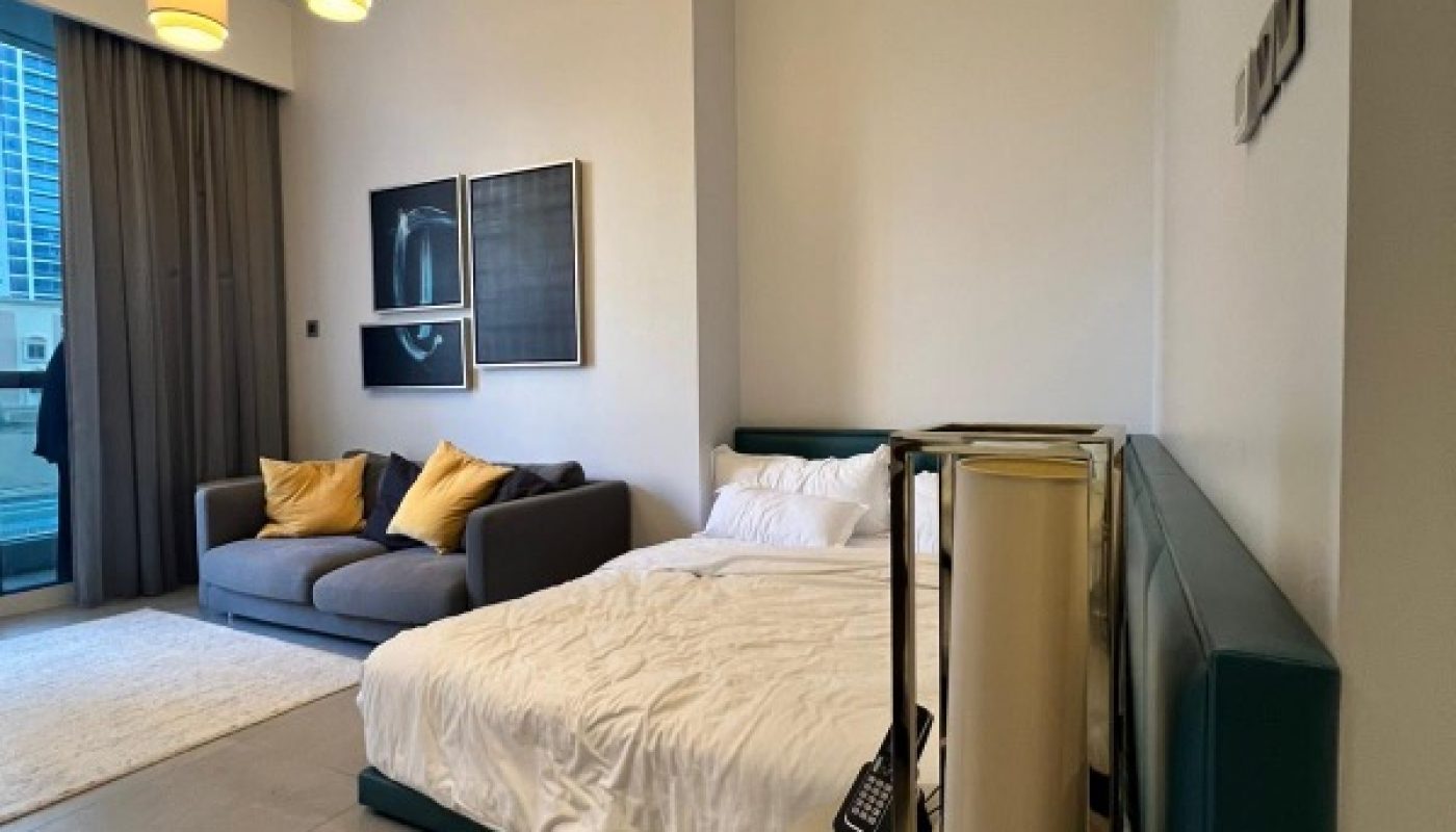 A modern studio room with a bed, gray sofa with yellow pillows, two pendant lights, wall art, and a round bedside table, with floor-to-ceiling windows and tiled flooring.