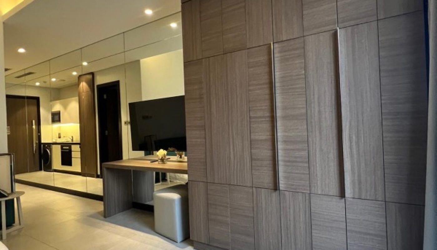 Modern hotel room with large wooden wardrobe, mirrored wall, desk, television, and kitchenette visible in the background.