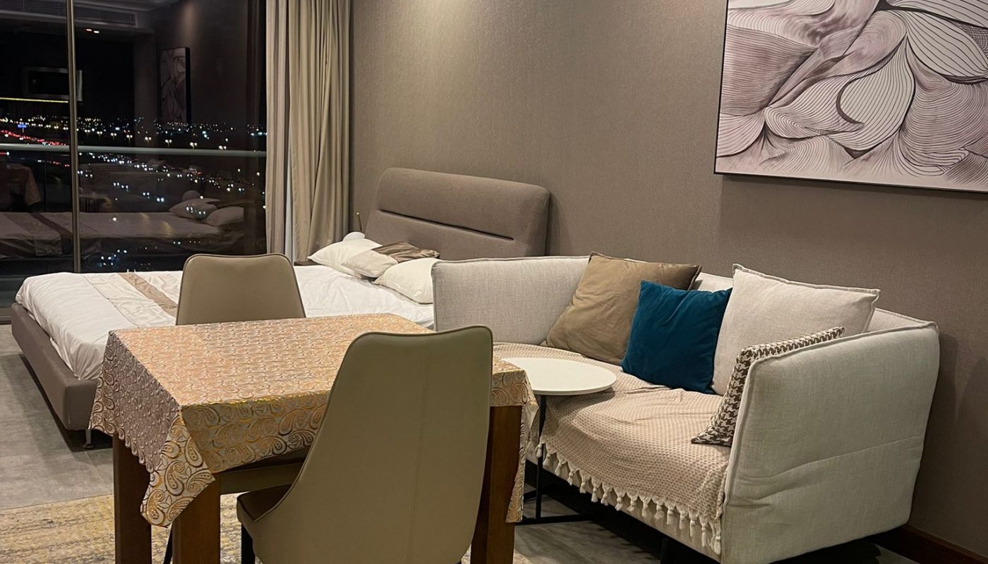 A modern living room with a beige sofa, a table with a lace cover, two chairs, a bed, wall art, and a large window showing city lights at night.