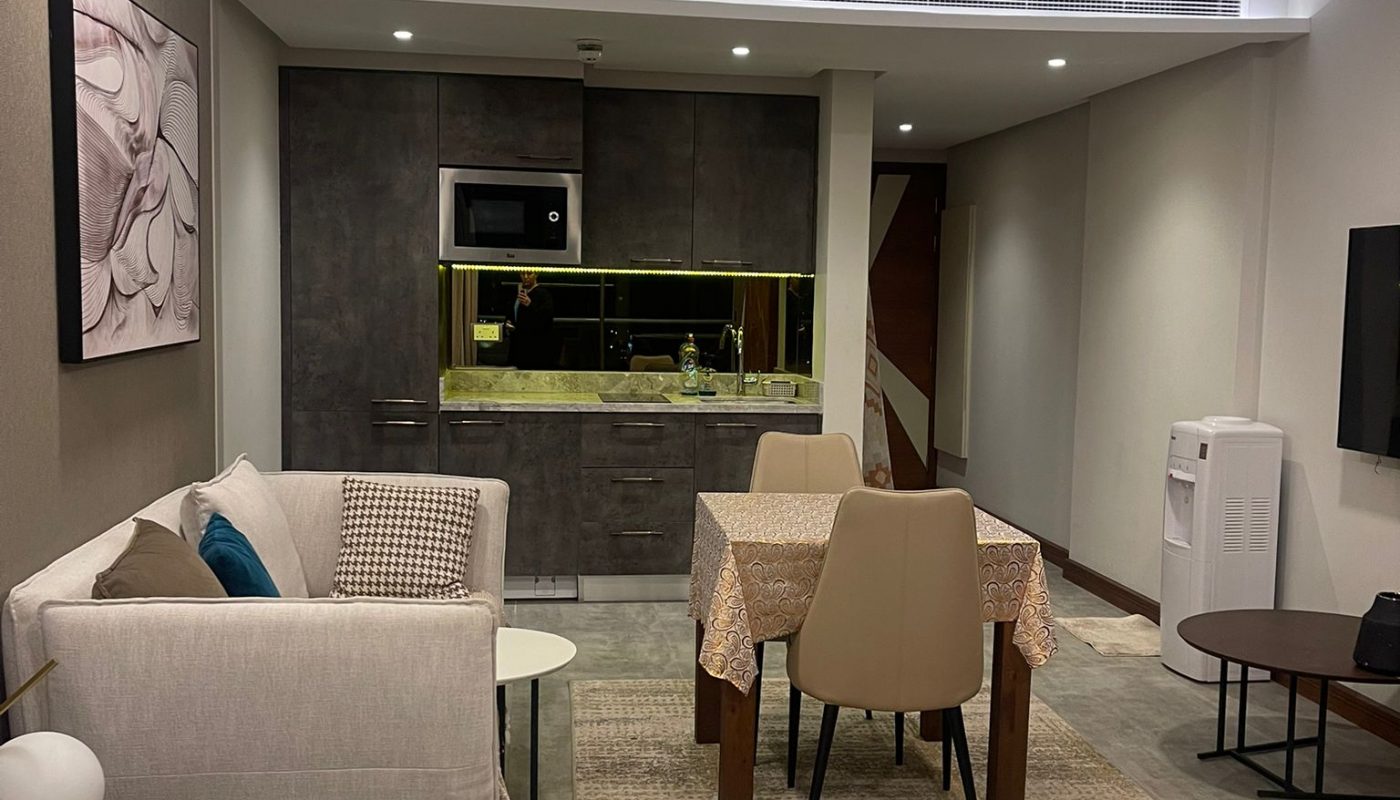 Modern studio apartment with bed, sofa, dining table, kitchenette, wall-mounted TV, and neutral decor. Room is well-lit with recessed ceiling lights.