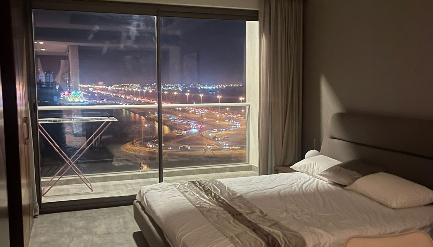 A modern bedroom with a neatly made bed, large glass doors opening to a balcony, and a night view of city lights and a highway outside.