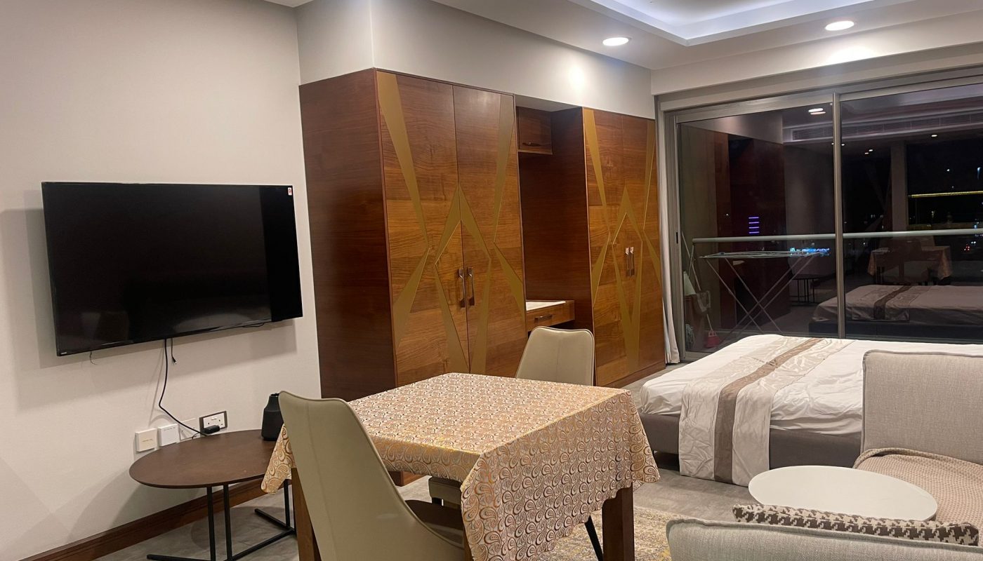 Modern studio apartment with a dining table, chairs, wall-mounted TV, bed, wardrobe, and a balcony visible through sliding glass doors at night.