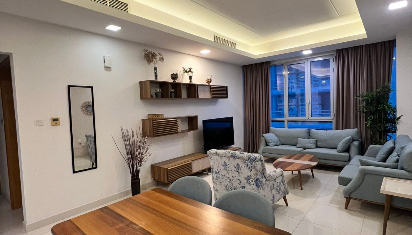 Modern living and dining area with wooden furniture, light blue chairs, a TV, shelves, and large window with curtains. Neutral color scheme and tiled floors.