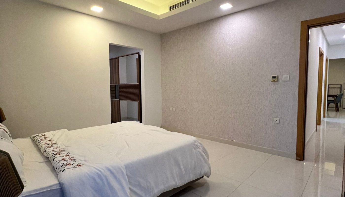 A modern bedroom with a double bed, white tile floor, light-colored walls, wooden nightstand, and a hallway visible through an open door.