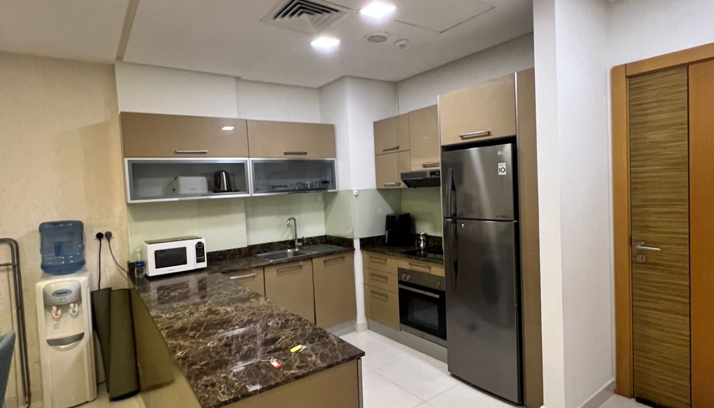 Modern kitchen with brown cabinets, marble countertop, stainless steel appliances, microwave, and a water dispenser on white tiled flooring. A closed wooden door is visible on the right.