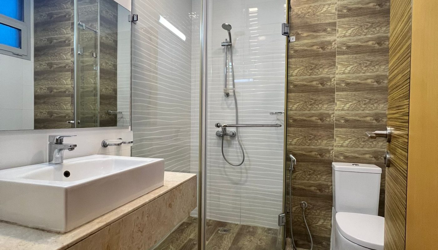 Modern bathroom with wood-patterned floor and wall, glass corner shower, white sink on stone countertop, and a toilet. Large mirror and ceiling lights are also visible.