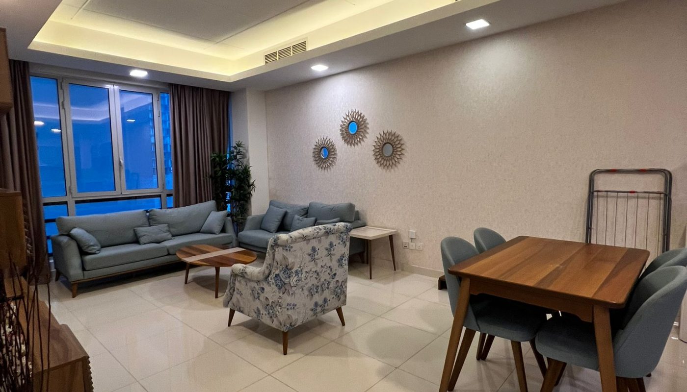 Modern living and dining area with tiled floors, light blue sofas, a patterned armchair, wooden dining table, and wall decor. Large windows with curtains provide natural light.