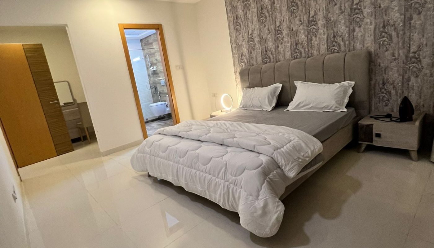 A modern bedroom with a double bed, white bedding, bedside tables, patterned accent wall, and an open door leading to a bathroom.