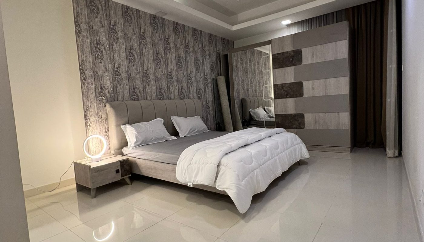 A modern bedroom with a large bed, patterned accent wall, nightstand with a circular lamp, floor mirror, wardrobe, white tile floor, and recessed ceiling lighting.