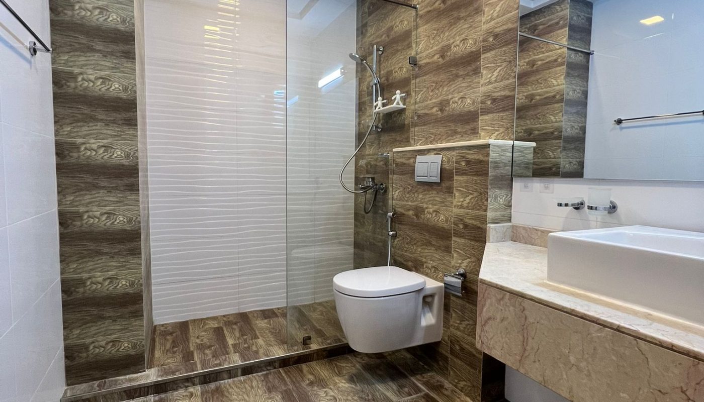 Modern bathroom with wood-patterned tiles, glass shower enclosure, wall-mounted toilet, marble countertop, and large mirror. Ceiling has recessed lights and vent.