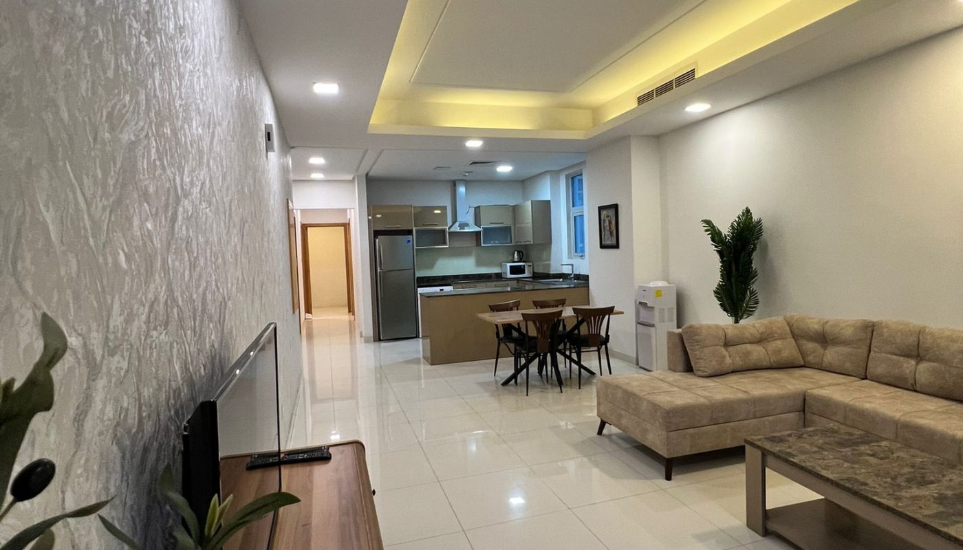 Modern open-plan living room and kitchen with white tiled floors, beige sectional sofa, wooden dining table, and recessed ceiling lighting.