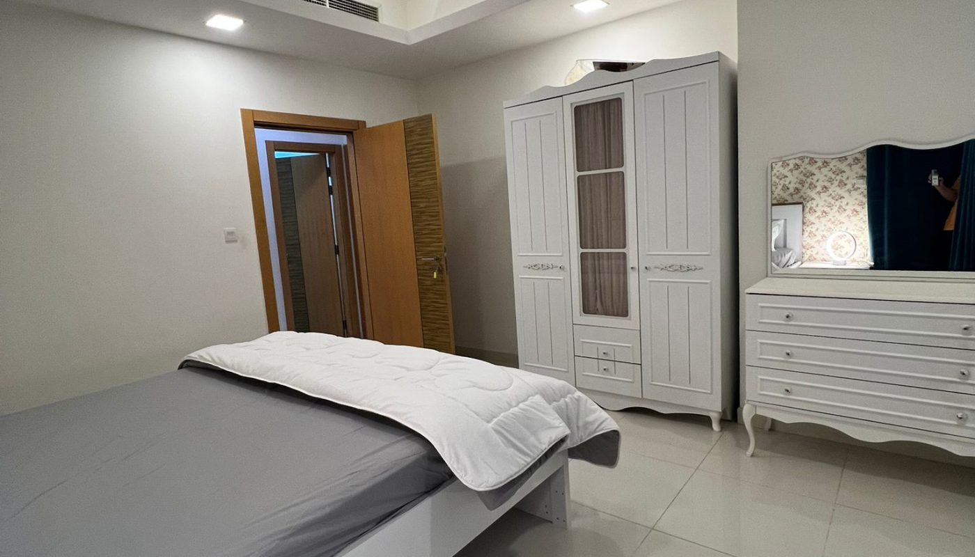 Minimalist bedroom with a gray-sheeted bed, white wardrobe, dresser with mirror, open wooden door, and tiled floor under recessed ceiling lights.