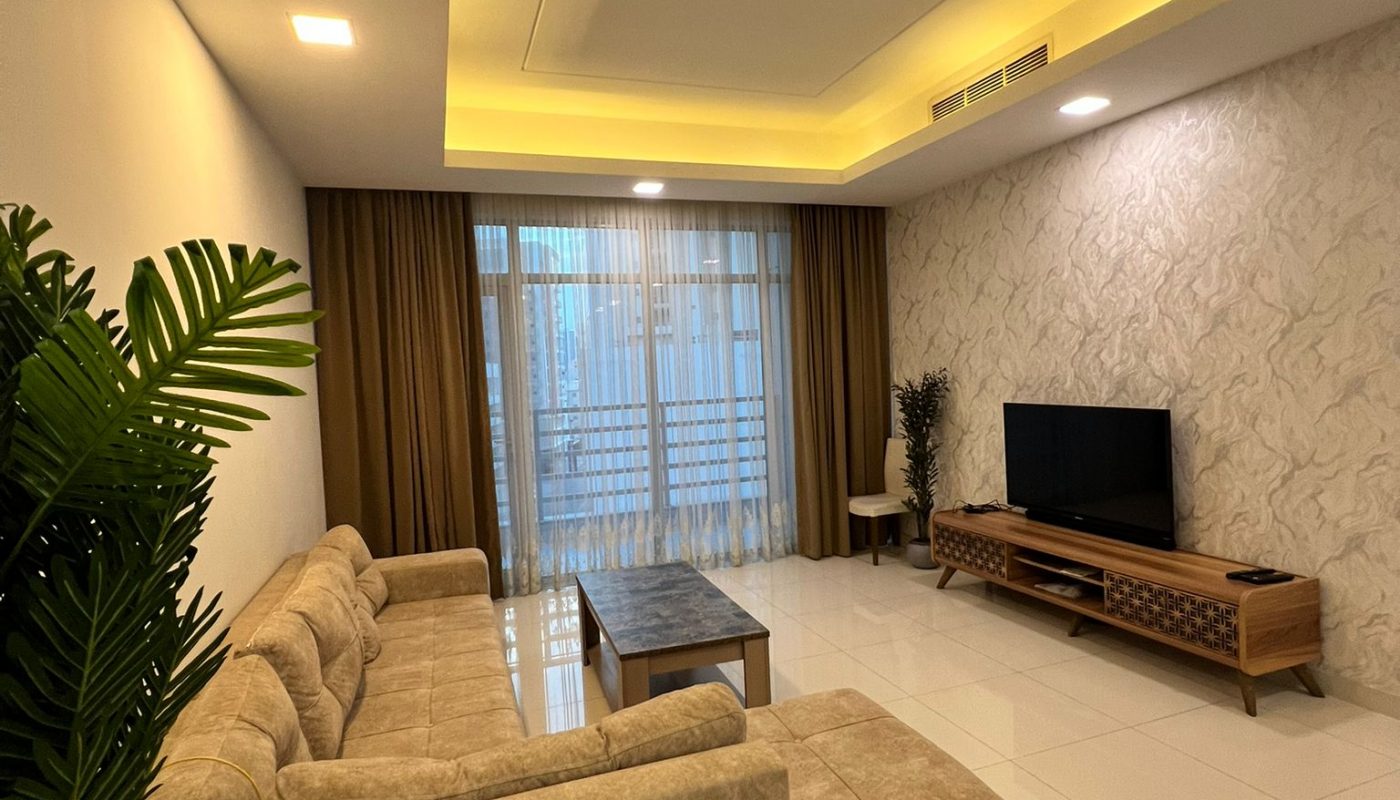 Modern living room with beige sectional sofa, coffee table, TV on a wooden stand, tiled floor, patterned wall, large window with curtains, and potted plants.