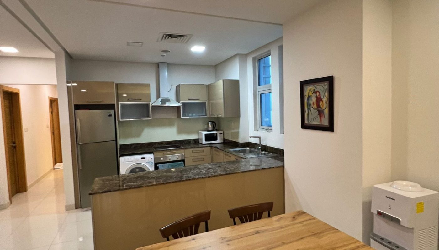 Modern kitchen and dining area with a wooden table, four chairs, marble countertop, stainless steel appliances, and wall art. Bright lighting and tiled floor.