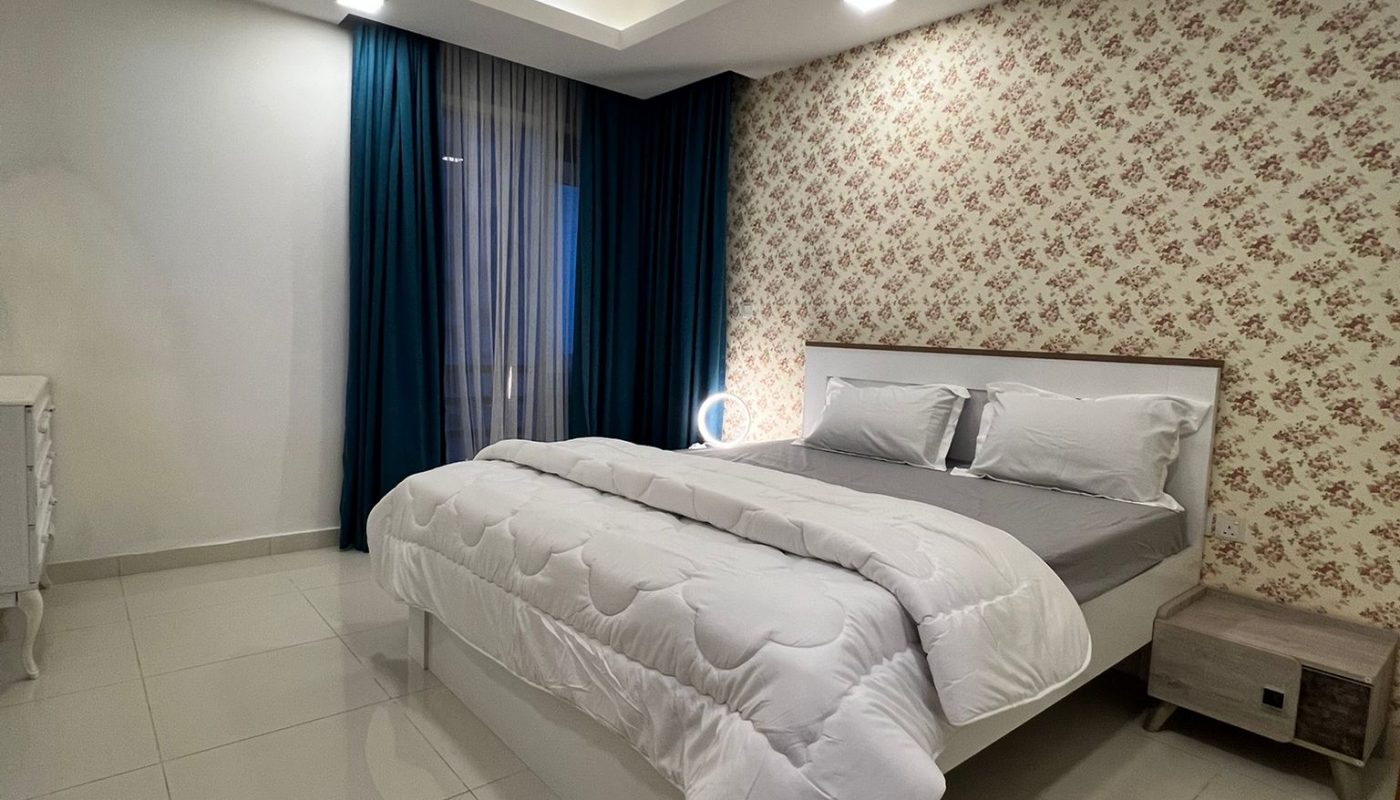 A neatly made bed with white bedding sits against a floral accent wall in a modern, tiled bedroom with blue curtains and minimal furniture.