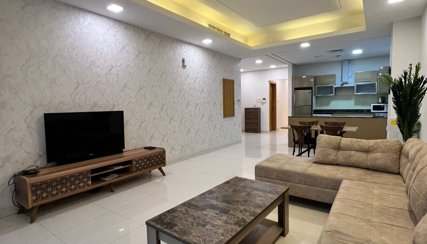 Modern living room with beige sectional sofa, marble coffee table, TV on a wooden stand, and an open-plan kitchen and dining area in the background.