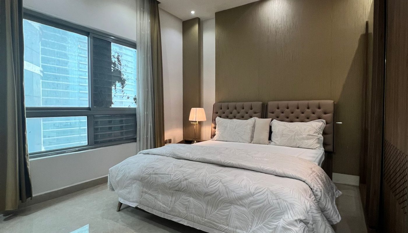 Modern bedroom with a large bed, white bedding, padded headboard, nightstand with lamp, floor-to-ceiling windows, and tiled floor.