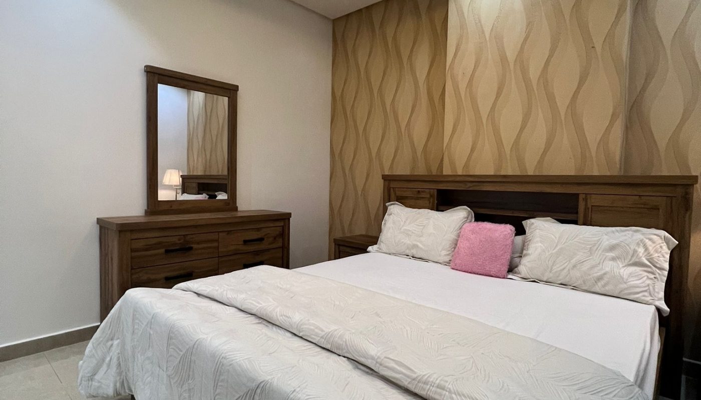 A neatly made bed with white bedding and a pink accent pillow in a modern bedroom with wood furniture, a dresser with a mirror, and patterned wallpaper.