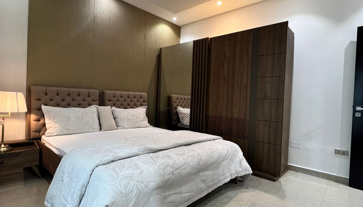 A modern bedroom with a double bed, white bedding, a bedside table with a lamp, a wooden wardrobe, tiled floor, and recessed ceiling lighting.