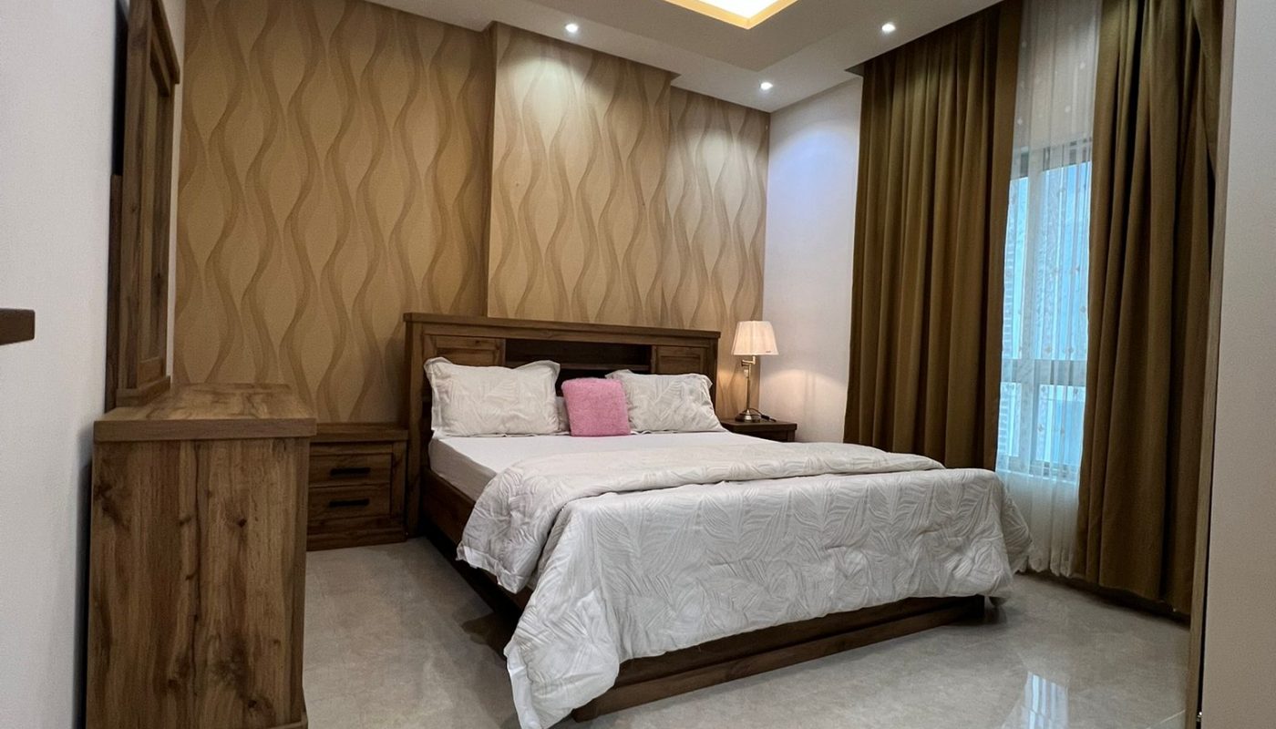 A modern bedroom with a wooden bed, white bedding, a dresser, bedside table with lamp, patterned accent wall, large window with curtains, and recessed ceiling lighting.