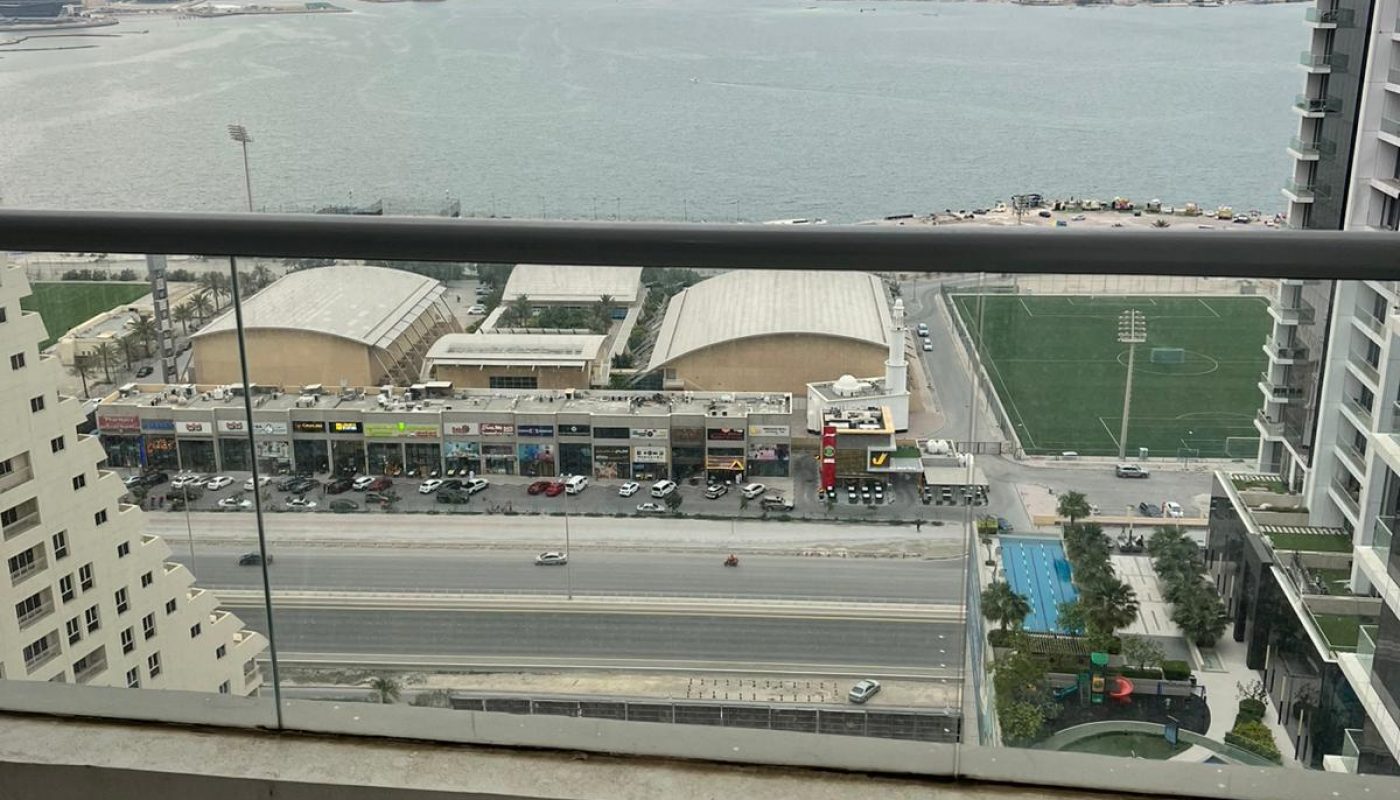 High-rise balcony view overlooking a busy road, sports field, and several buildings, with a body of water and distant shoreline visible under a cloudy sky.