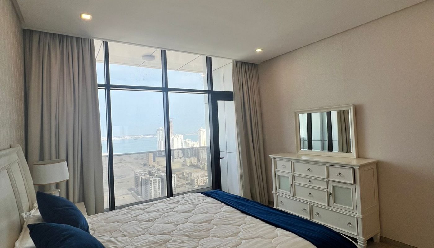 A modern bedroom with a large bed, white bedding, blue accents, dresser with mirror, floor-to-ceiling windows, and a city skyline view.