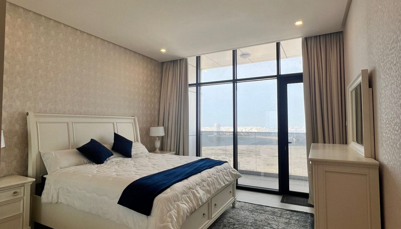 A modern bedroom with a large bed, white furniture, floor-to-ceiling windows, and a view of the outdoors.
