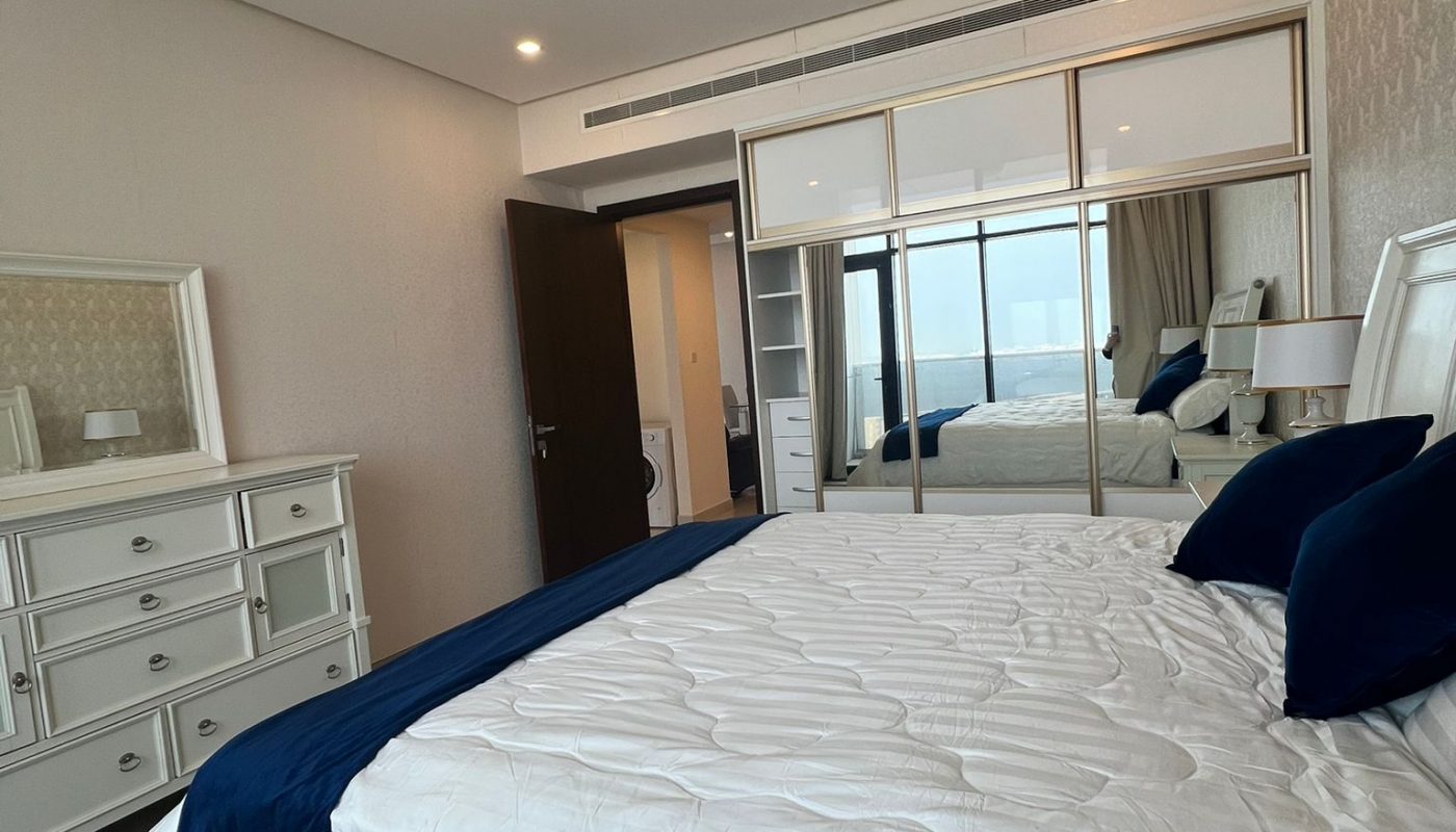 A modern bedroom with a white bed, navy blue bedding, a matching dresser, mirrored closet doors, and a window with city views.