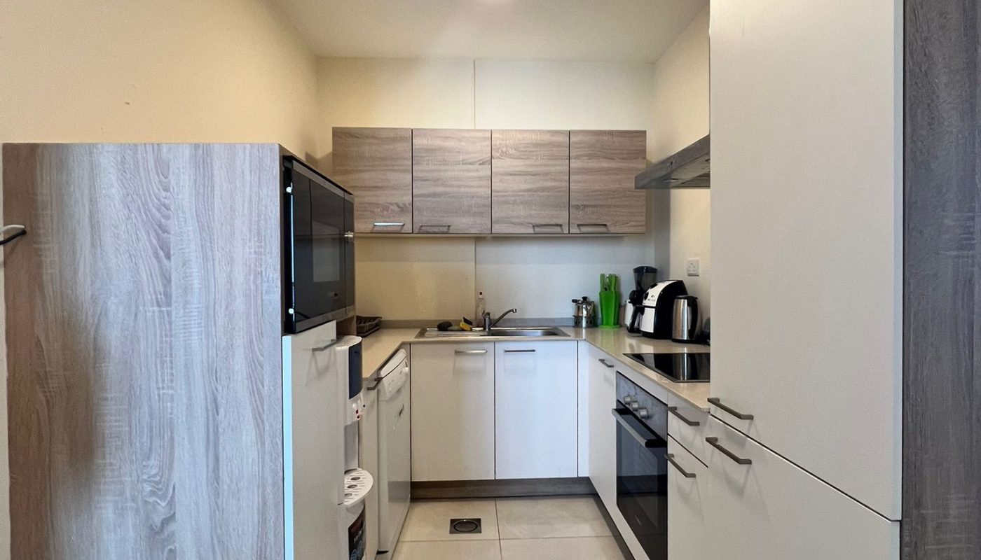 Modern kitchen with light wood and white cabinets, appliances including an oven, microwave, kettle, and toaster, and a sink. Trash bin visible in the foreground.